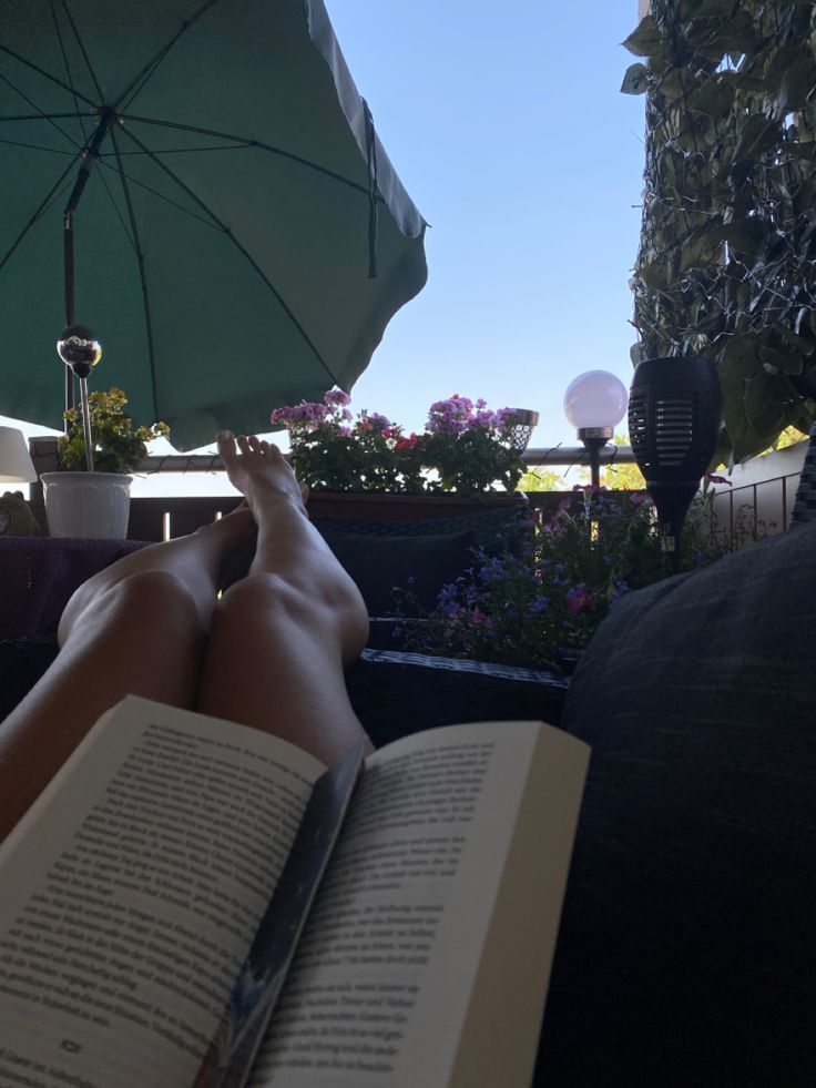 a person laying down reading a book with an umbrella over their head and flowers in the background