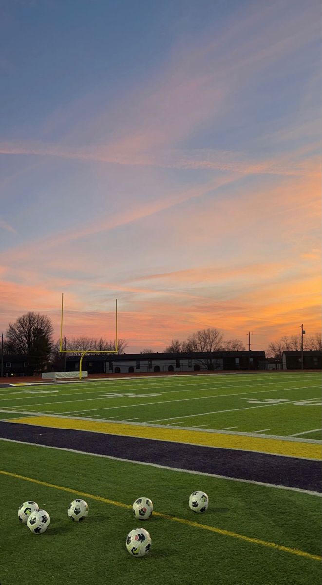 several soccer balls sitting on the grass in front of an empty football field at sunset