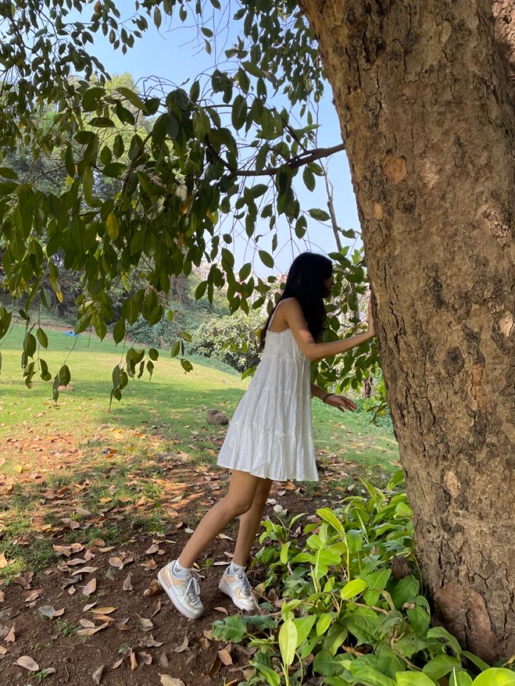 a woman in a white dress leaning against a tree with her hand on the trunk