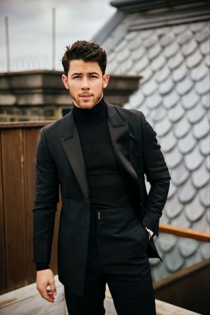 a man in a black suit and turtle neck sweater standing on a roof with his hands in his pockets