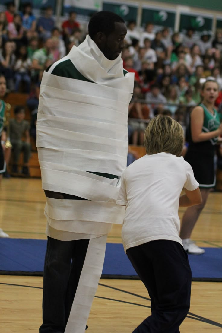 two people in white shirts and black pants standing on a basketball court with a giant piece of paper wrapped around them