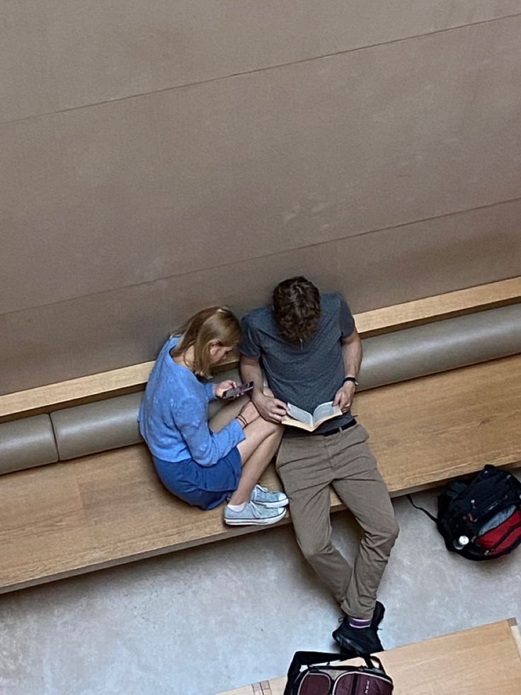 two people are sitting on a bench with their luggage and looking down at the floor