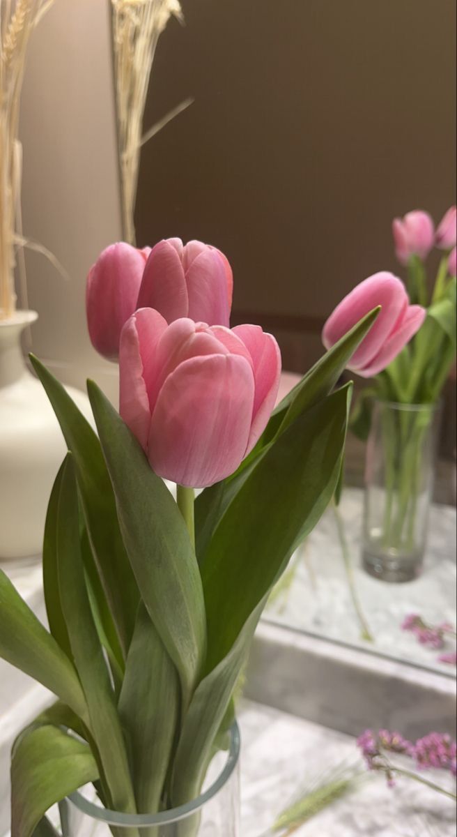 pink tulips are in a clear vase on a table with other glass vases