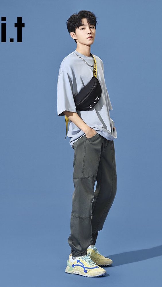 a young man standing in front of a blue background wearing a gray shirt and black pants