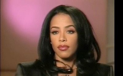 a close up of a person with long hair and wearing a black jacket, sitting in front of a pink wall