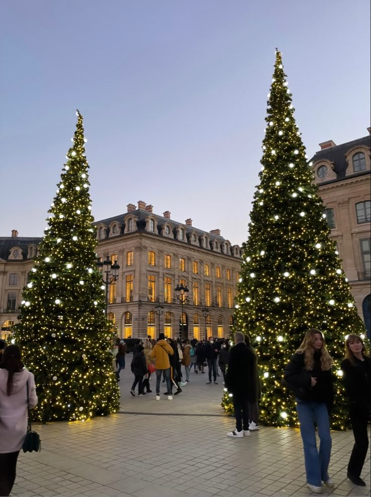 people are walking around christmas trees in front of a large building with lights on it