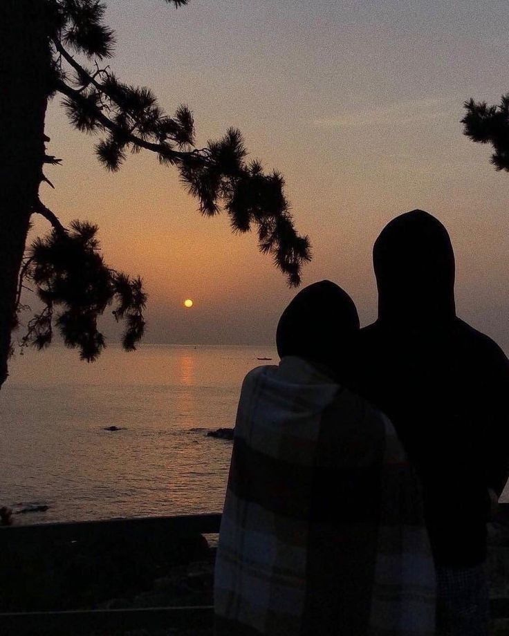 two people sitting on a bench watching the sun go down over the water at dusk