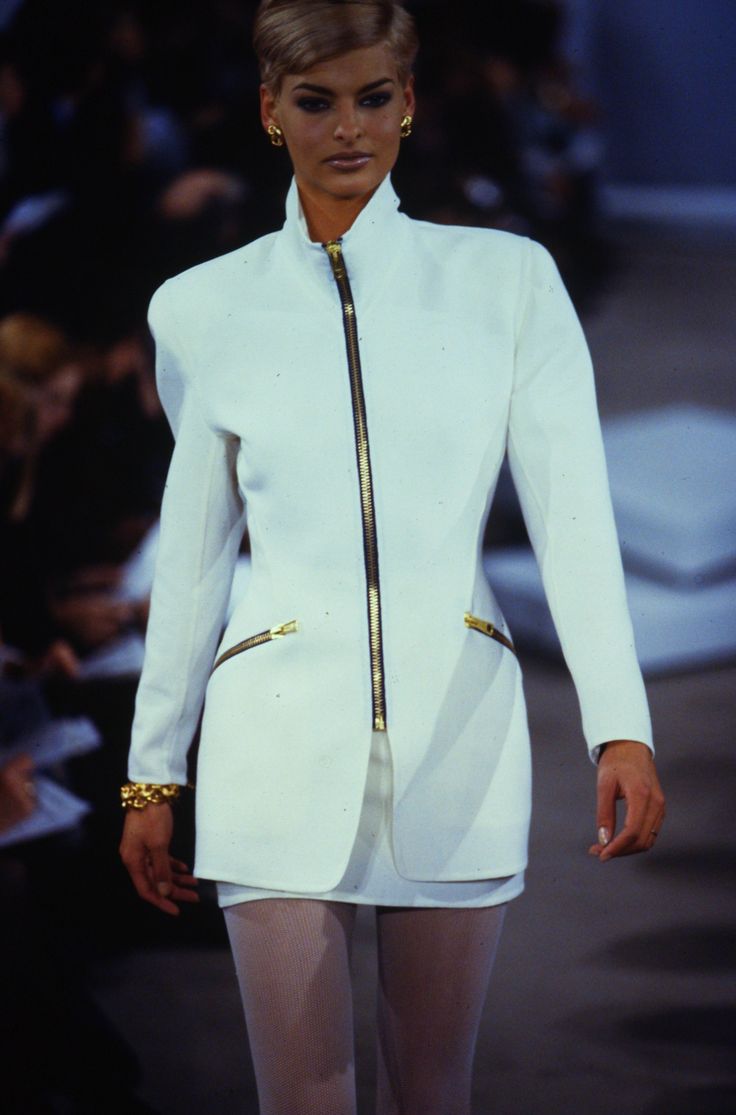 a model walks down the runway wearing white clothes with gold accents on her shoulders and legs