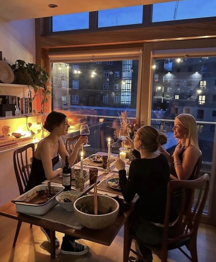 three women sitting at a table with food and drinks in front of a large window