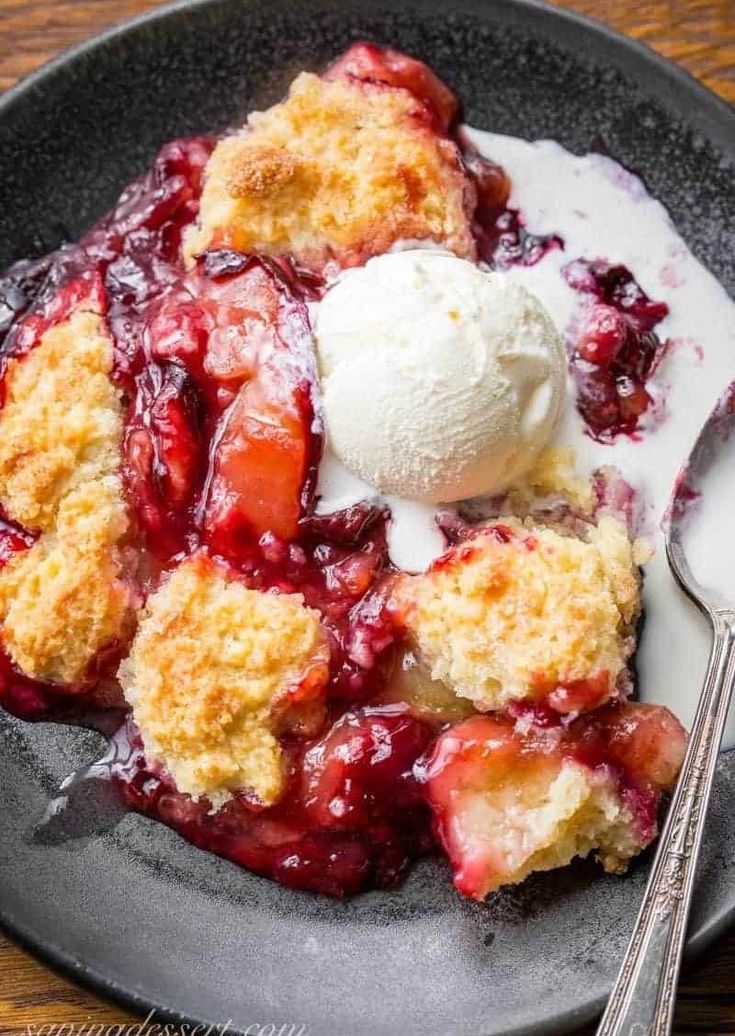 a black plate topped with fruit cobbler and ice cream on top of a wooden table