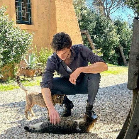 a man kneeling down petting a cat on top of a gravel ground next to a building