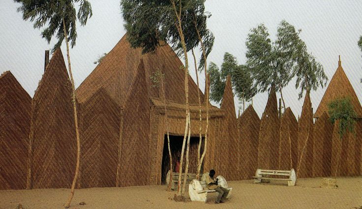 a man sitting on a bench in front of a building made out of sticks and palm trees