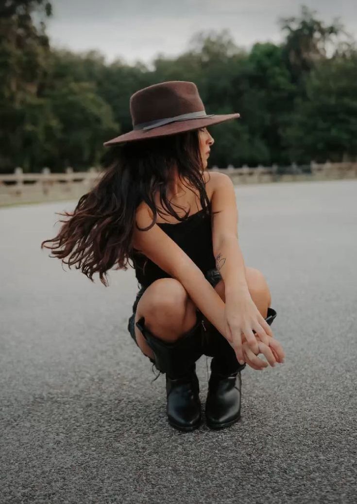 a woman kneeling down in the middle of an empty parking lot wearing a hat and boots