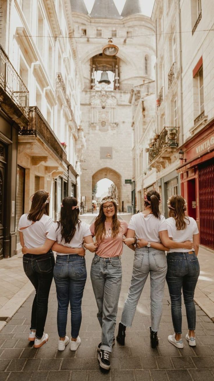 four girls are walking down the street in jeans and t - shirts with their arms around each other