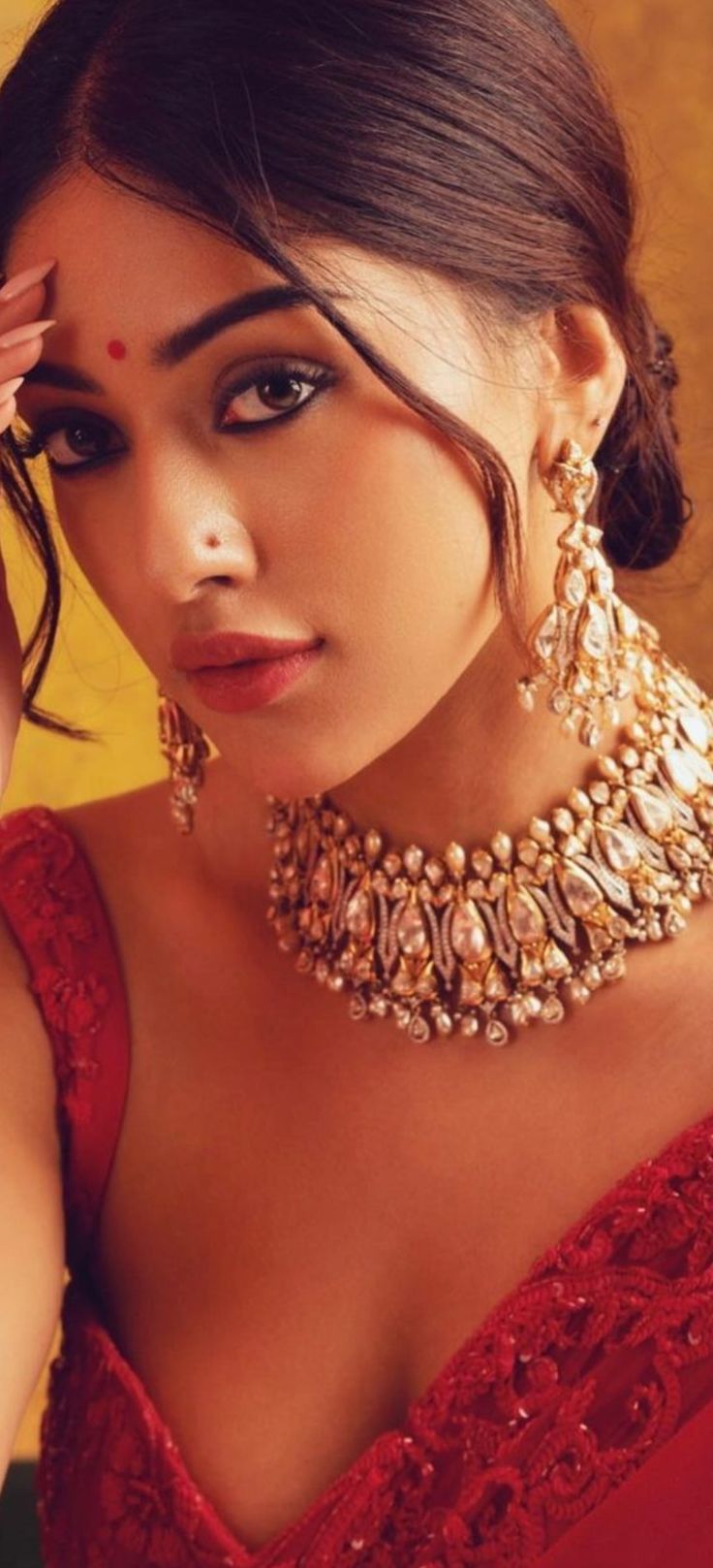 a beautiful woman in a red dress posing with her hand on her head and wearing gold jewelry