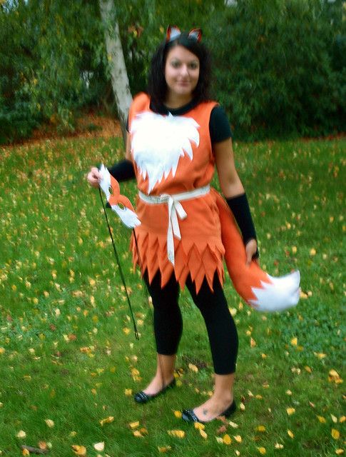 a woman dressed in an orange and white fox costume standing on grass holding a stick