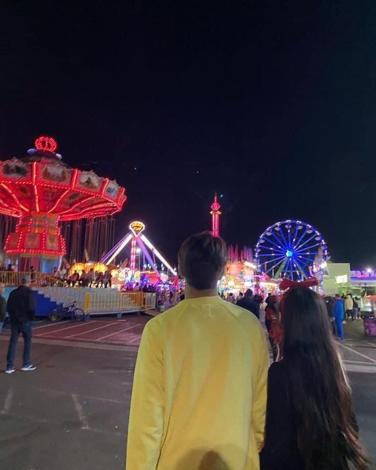 two people are standing in front of an amusement park at night with ferris wheel and carnival rides