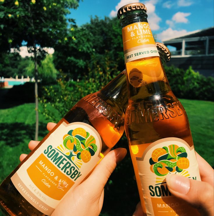 two bottles of beer are being held by someone's hands in front of some trees