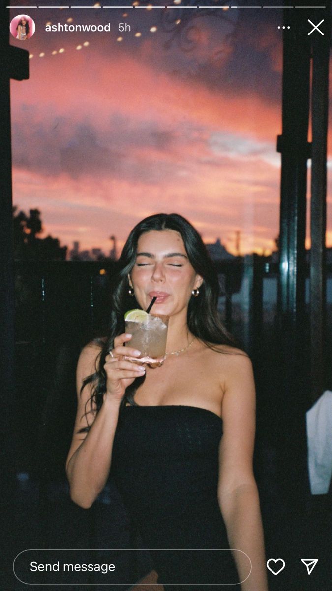 a woman in a strapless dress is drinking from a drink glass at sunset with the sky behind her