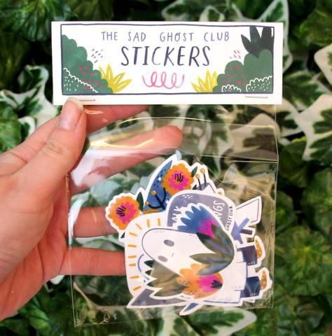 a person holding up a sticker in front of some leaves and plants with the words, the lad ghost club stickers below it