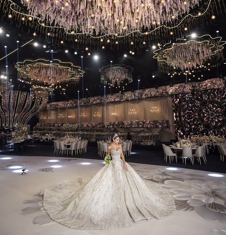 a woman in a wedding dress standing under chandelier at a banquet hall with tables and chairs