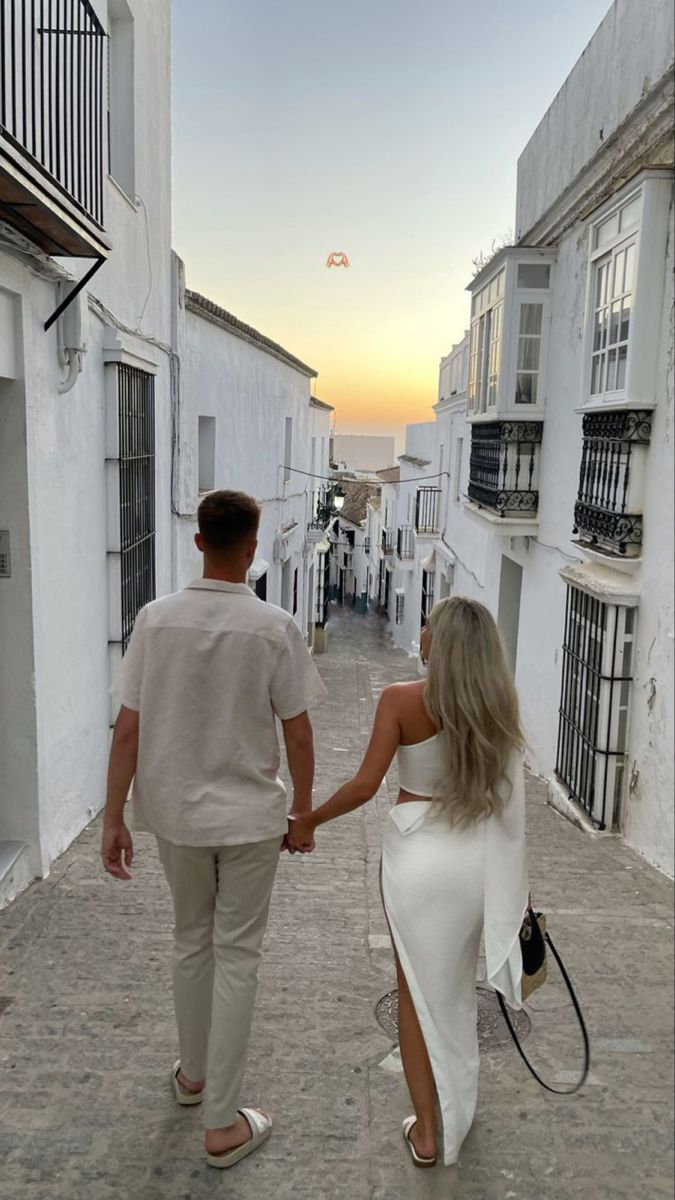 a man and woman holding hands walking down an alley way with buildings in the background