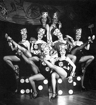 a black and white photo of women in costumes with dice on their heads posing for the camera