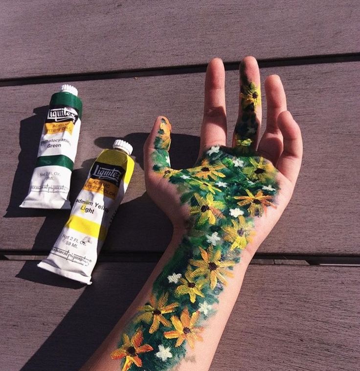 a person's hand painted with flowers and leaves next to some paint tubes on a wooden surface