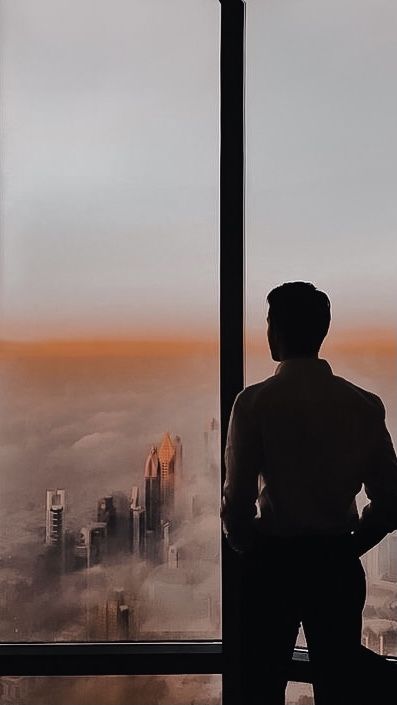 a man standing in front of a window looking out at the city below and skyscrapers