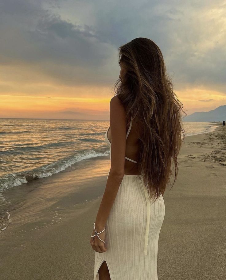 a woman standing on top of a sandy beach next to the ocean with her back to the camera