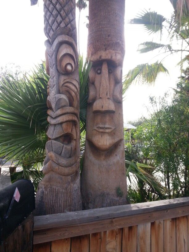 two carved totems sitting next to each other on a wooden fence in front of palm trees