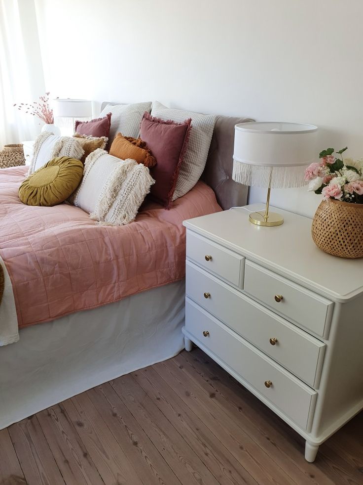 a white bed with pink and gold pillows on top of it next to a dresser