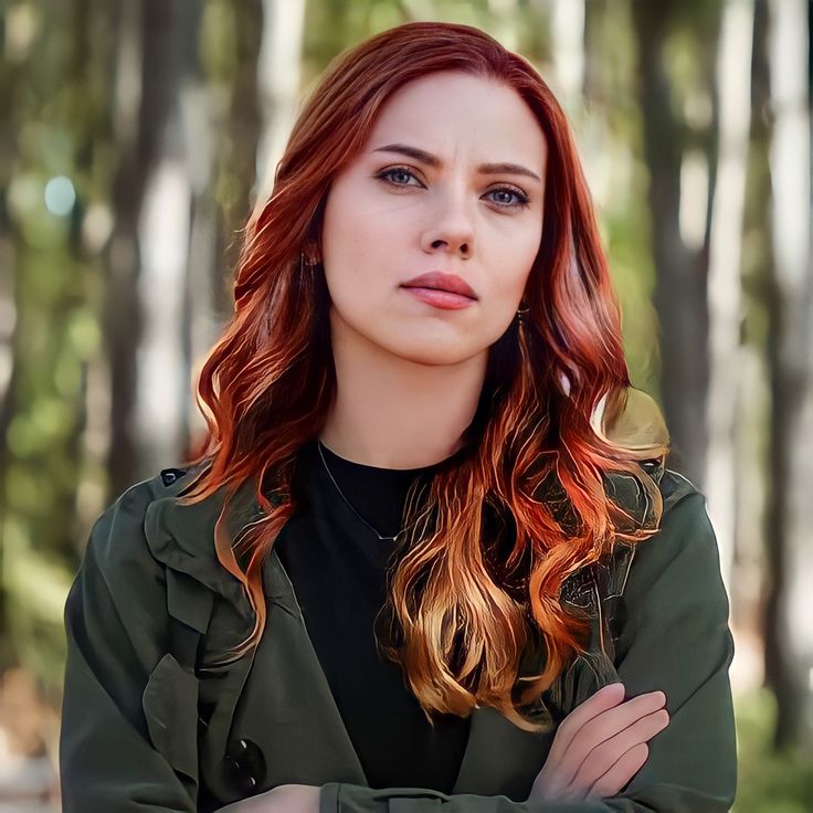 a woman with red hair is posing for a photo in front of some trees and bushes