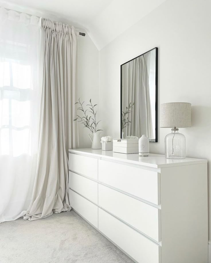 a white dresser with a mirror and vase on it in front of a window covered by drapes