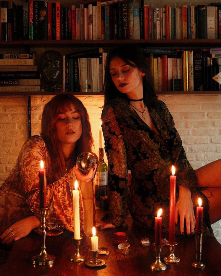 two women sitting at a table with candles in front of them and books on the shelves behind them