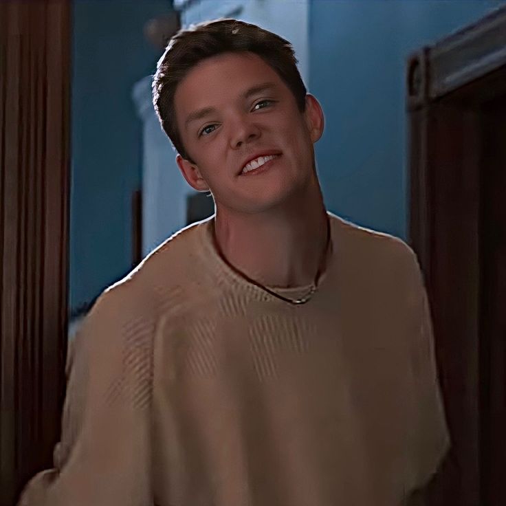 a young man in a tan sweater smiles at the camera while standing next to a doorway