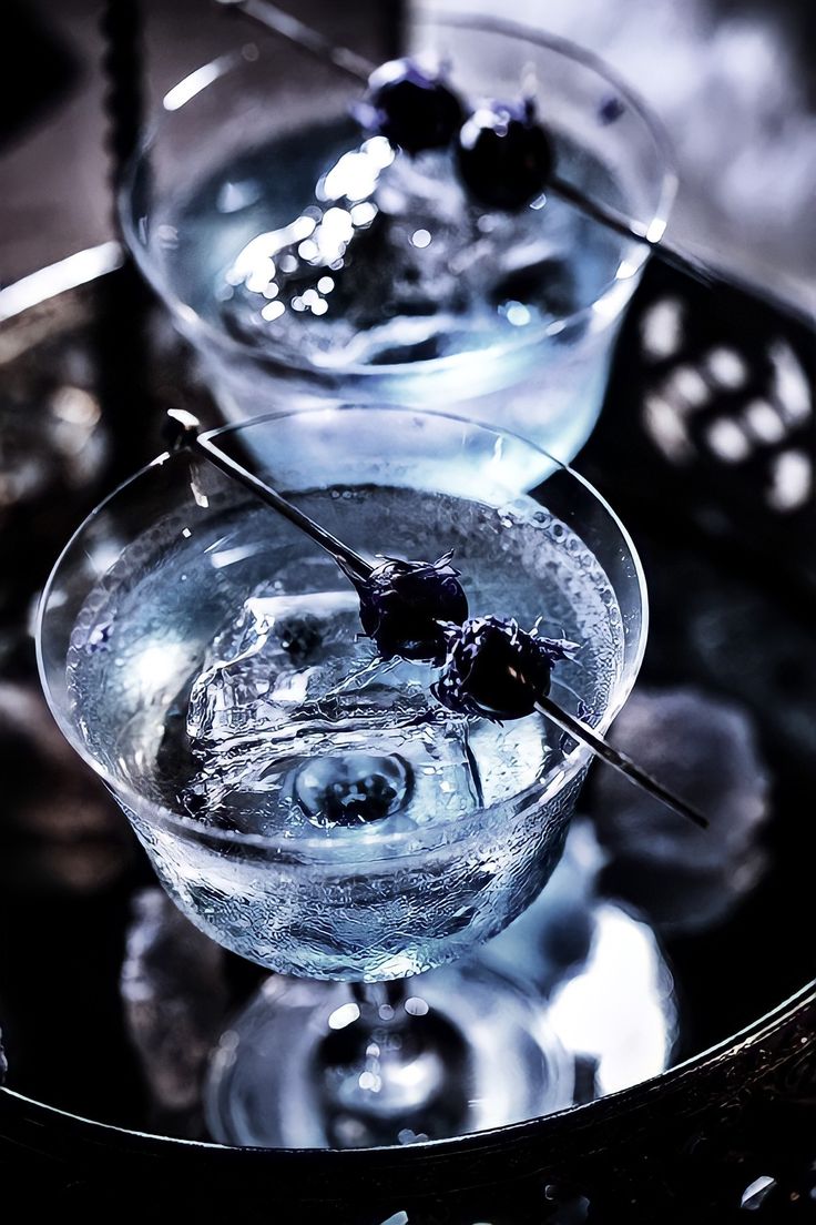 two glasses filled with water and some black olives on top of each glass bowl