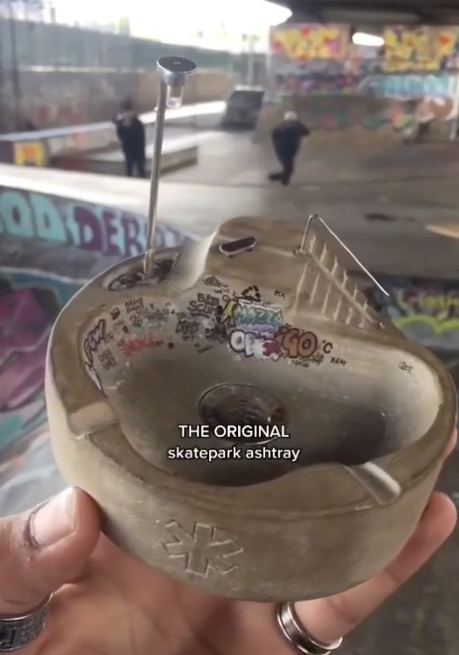 a person holding up a skateboard in front of a graffiti covered wall with the words, the original skeeeppark ashtrayy on it