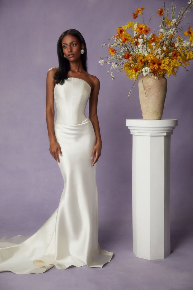 a woman standing next to a vase with flowers in it and wearing a white dress