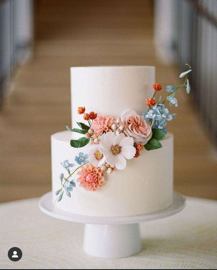 a three tiered white cake with pink and blue flowers on the top is sitting on a table