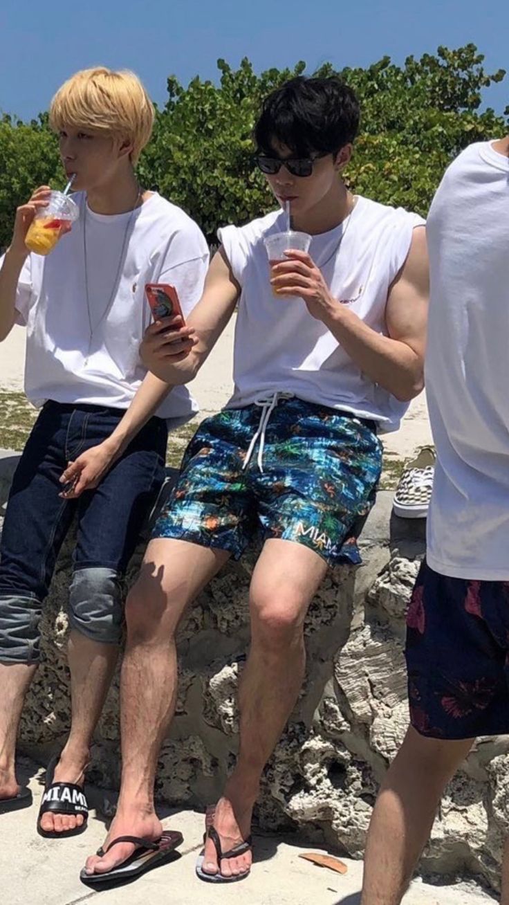 three young men sitting next to each other on a stone wall drinking drinks and looking at their cell phones