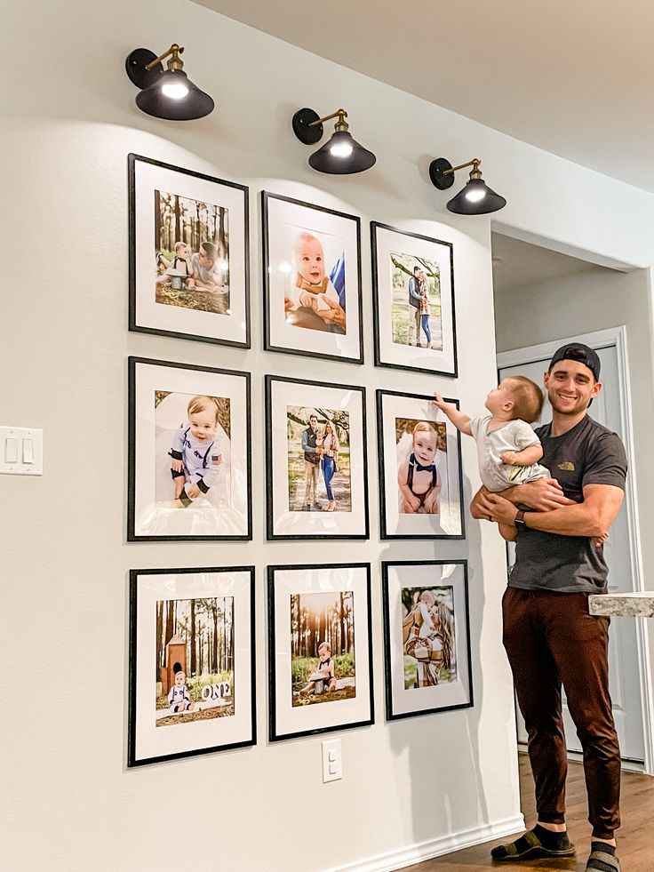 a man holding a baby in front of a wall with pictures hanging on the wall