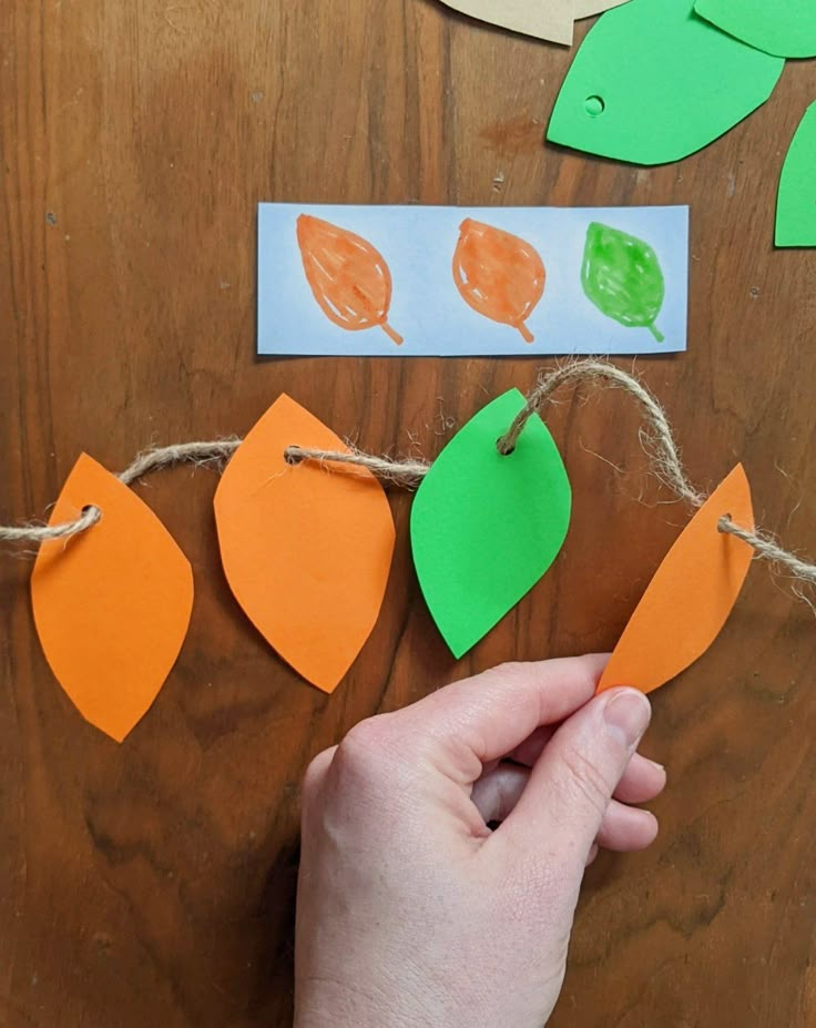 a hand is holding an orange piece of paper and string with green leaves on it