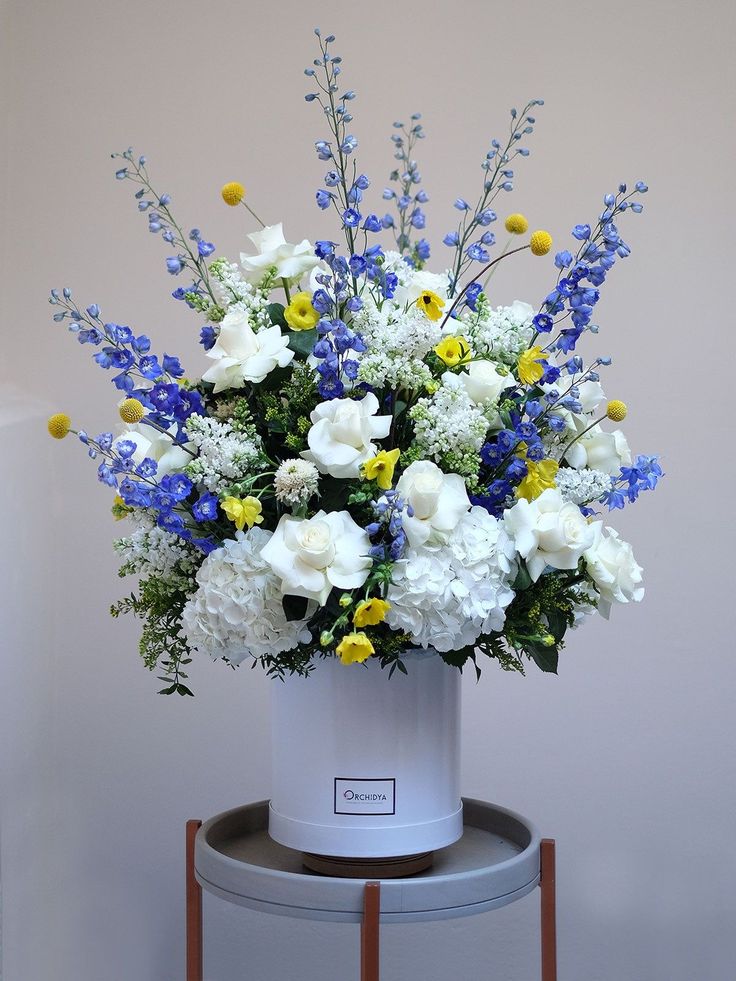 a white vase with blue, yellow and white flowers sitting on top of a table