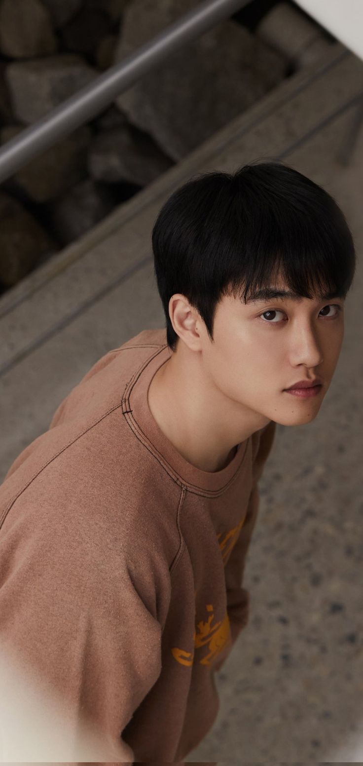 a young man with black hair and brown shirt looking at the camera while standing in front of some rocks