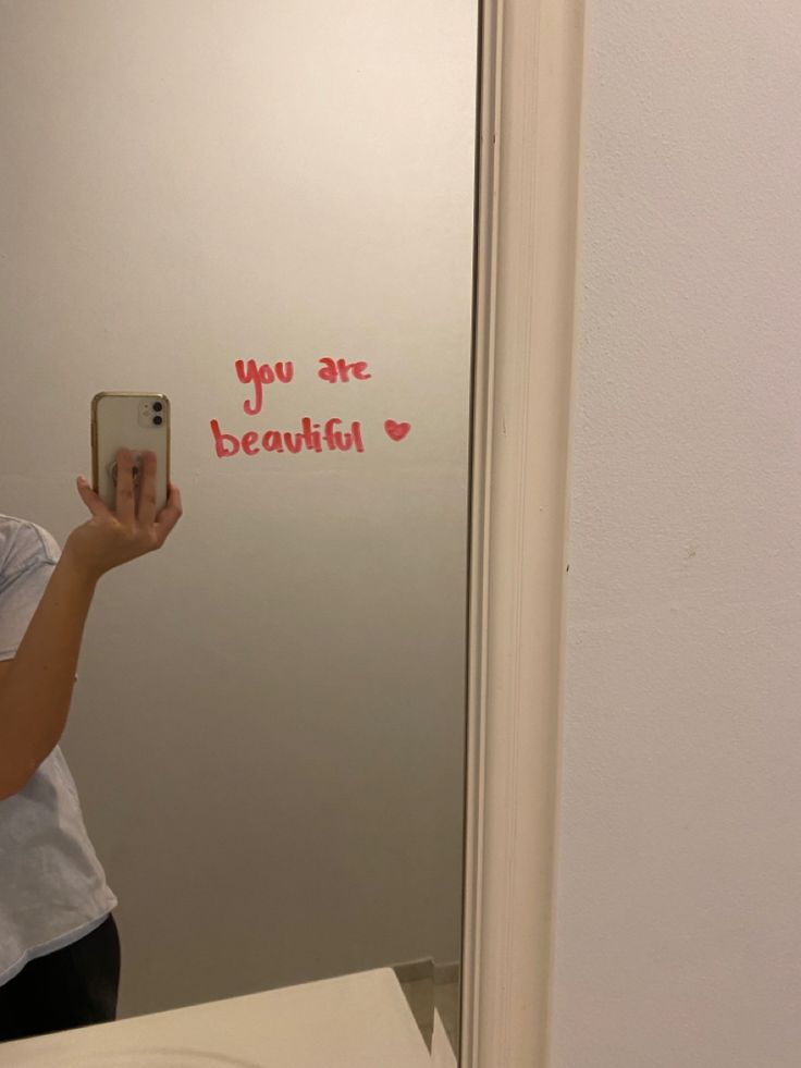 a woman taking a selfie in front of a mirror with the words you are beautiful written on it