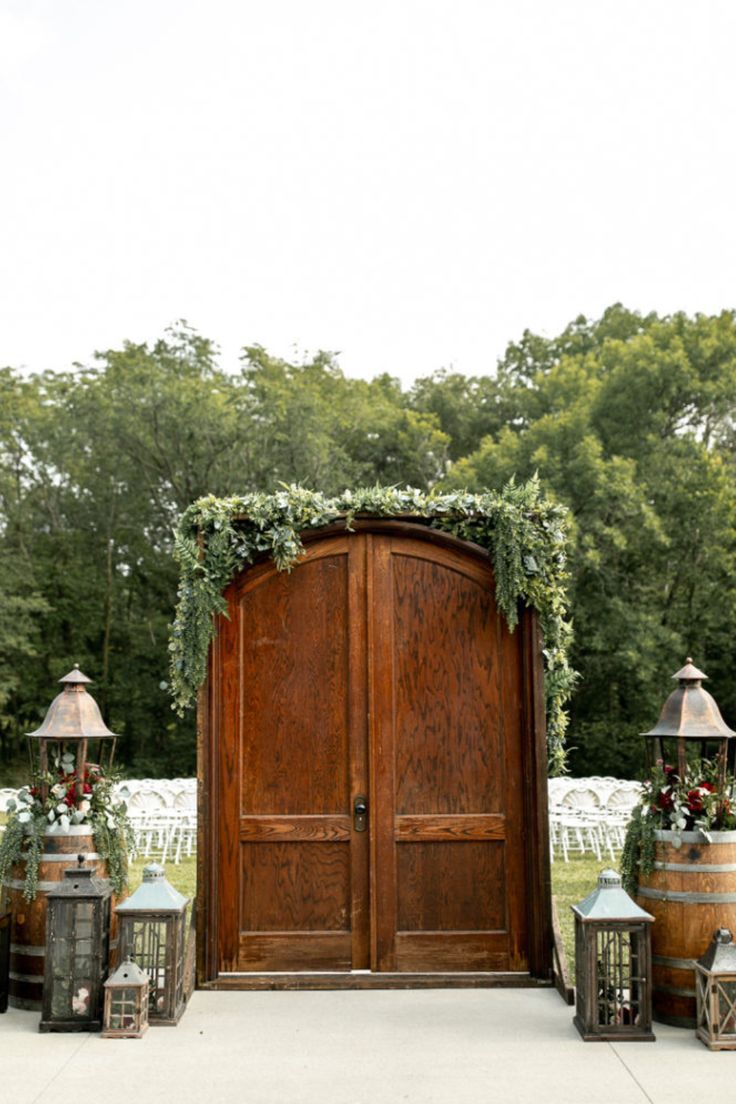 Enchanting outdoor wedding entrance to ceremony with beautiful fall florals and lanterns surrounding gorgeous wooden doors. Wedding Doors Outside Outdoor Ceremony, Outdoor Ceremony Entrance, Outdoor Ceremony Entrance Ideas, Wedding Door Entrance Outside, Wedding Outdoor Doors, Outdoor Wedding With Doors Entrance, Wedding Entrance Outdoor Ceremony, Outside Wedding Doors Entrance, Wedding Ceremony Doors Entrance