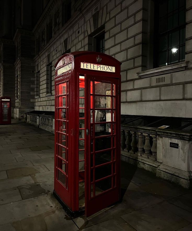 a red phone booth sitting in the middle of a street at night time with its lights on