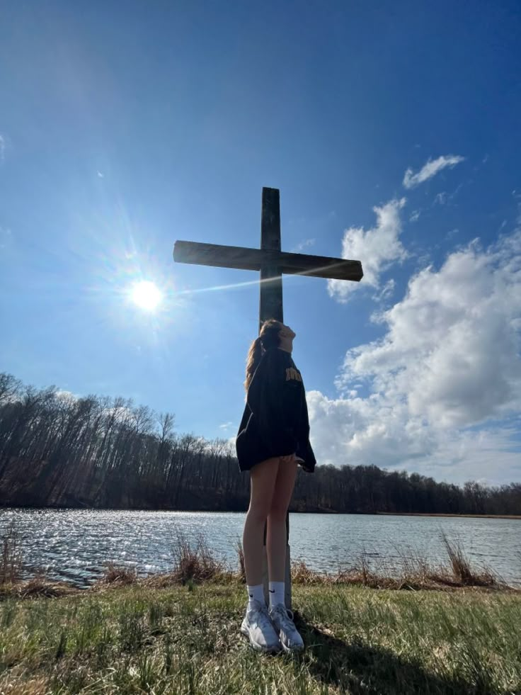 a woman standing in front of a cross on top of a field next to a lake