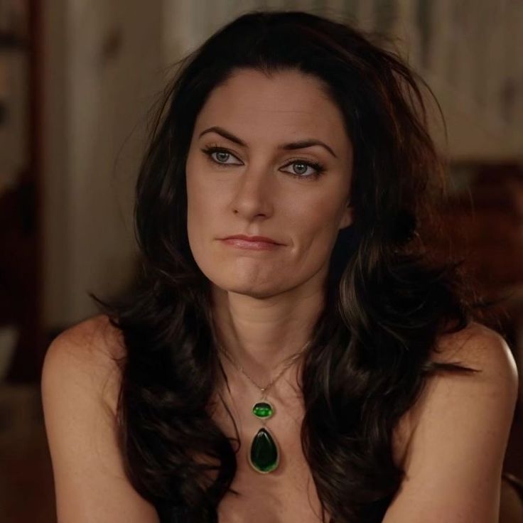 a woman with long dark hair wearing a green necklace and sitting in front of a couch